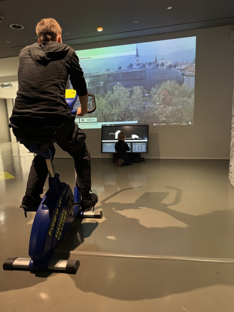 Mathias Kessler, Pump it Up!, visitor pedaling the stationary bike with the live Kunsthaus Graz square webcam projected behind, Kunsthaus Graz, 2024.
